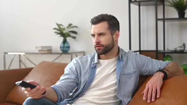Egy fiatal szakállas férfi kezében TV távirányítóval, TV-t néz — Stock videók