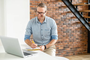 Genç çalışkan bir çalışan masada oturuyor ve dizüstü bilgisayara bakarken bir şeyler yazıyor.