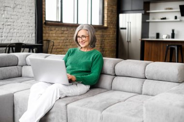 Evde dizüstü bilgisayar kullanan olgun bir yaşlı kadın.