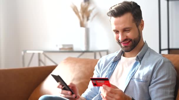 Cheerful young man holds credit card and a smartphone in hands, shopping online — Stock Video