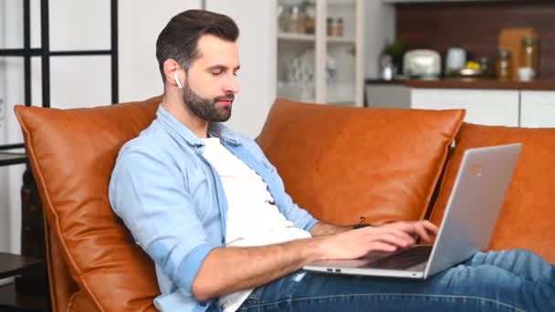 Un hombre guapo con camisa casual usando un portátil sentado en casa — Vídeos de Stock