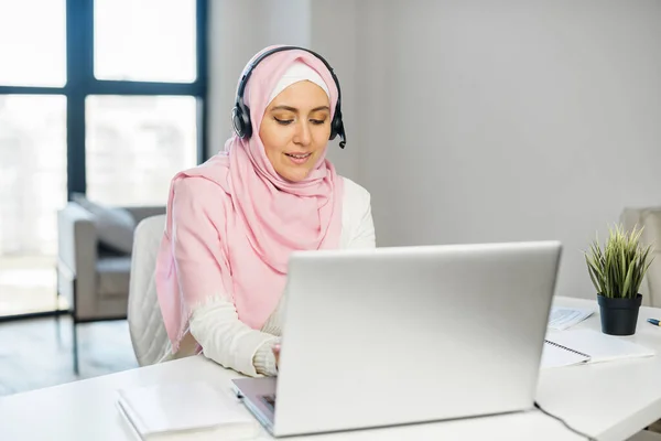 A young muslim woman wearing hijab using a laptop and a headset