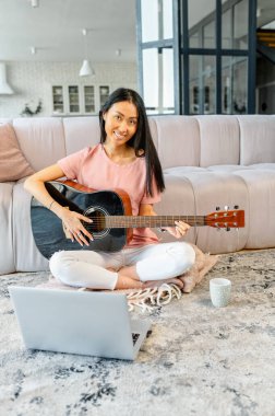 Çinli bir kadın dizüstü bilgisayarının önünde gitarıyla yerde oturuyor.