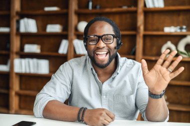 Siyahi, dost canlısı, kulaklık takmış, el sallıyor, içeride oturuyor.