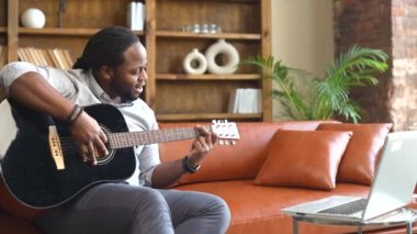 Dizüstü bilgisayarın önünde gitar çalan Afro-Amerikalı bir adam.