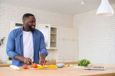 Laptopta video dersi izlerken konsantre olmuş Afrikalı bir adam yemek pişiriyor.