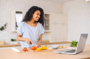 Laptopta yüksek lisans yaparken gülümseyen melez bir kız yemek pişiriyor.
