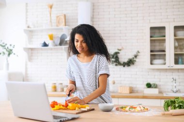 Laptopta yüksek lisans yaparken gülümseyen melez bir kız yemek pişiriyor.