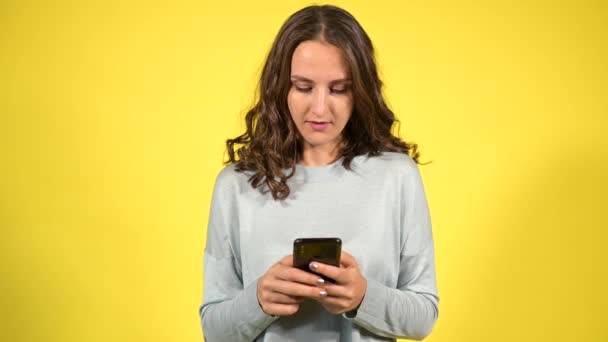 Ελκυστική σγουρή νεαρή γυναίκα χρησιμοποιώντας smartphone απομονωμένο σε κίτρινο — Αρχείο Βίντεο