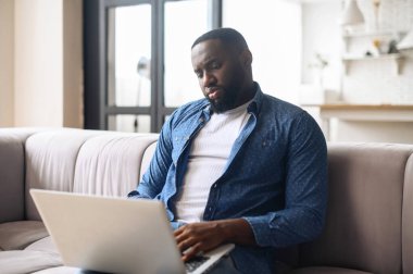 Kanepede oturan siyah adam dizüstü bilgisayar kullanıyor.