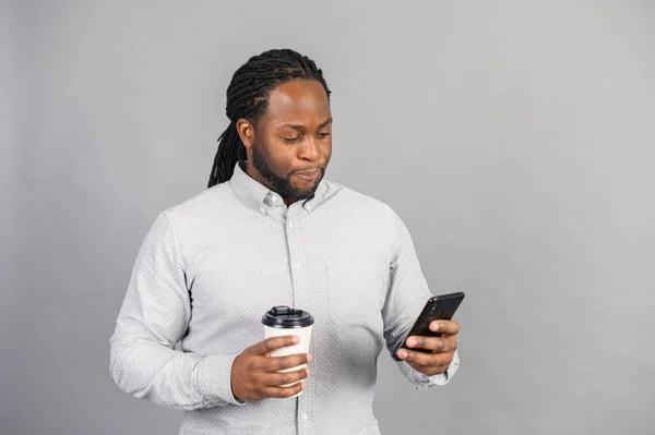 Sonriente hombre afroamericano sosteniendo teléfono y cofee, aislado en gris — Foto de Stock