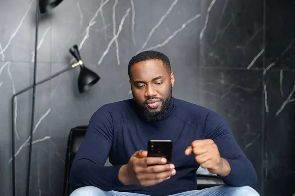 Koncentrerad afrikansk man som sitter på soffan och tittar på mobilskärmen — Stockfoto