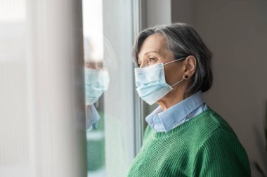 Yüzünde maske olan yaşlı bayan pencereye bakıyor.
