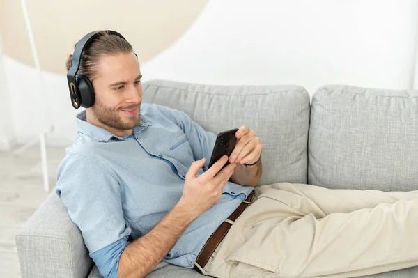 Chico guapo satisfecho usando smartphone y auriculares inalámbricos para escuchar música —  Fotos de Stock