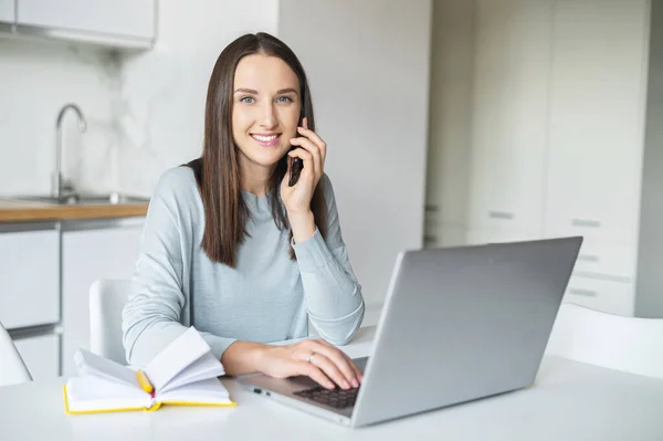 Gülümseyen ve pozitif genç kadın bilgisayarla çalışırken akıllı telefondan konuşuyor. — Stok fotoğraf