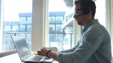 Kendine güvenen bir erkek işçi elinde dizüstü bilgisayarla masada oturuyor ve uzaktan çalışıyor.