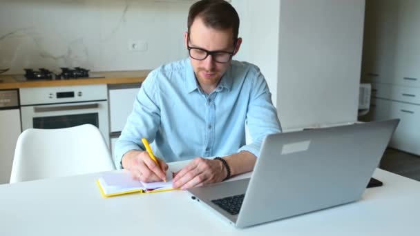 Fokuserad manlig entreprenör anteckningar med hjälp av en bärbar dator för distansarbete hemifrån — Stockvideo