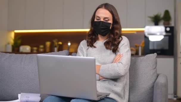 En ung kvinna med skyddande ansiktsmask sitter på soffan med en laptop — Stockvideo