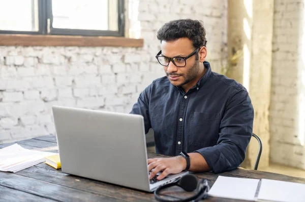 Fokuserad hindu frilansare kille bär glasögon med bärbar dator i samtida kontorsutrymme — Stockfoto