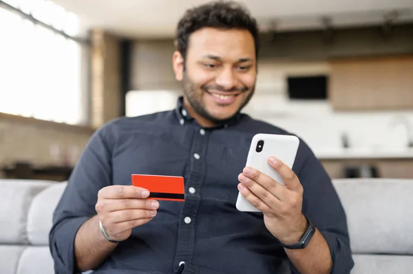 Homem índio sorridente detém cartão bancário e smartphone — Fotografia de Stock