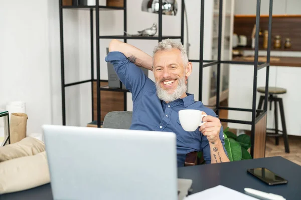 Fröhlich reifer männlicher Mitarbeiter genießt Morgenkaffee am Arbeitsplatz im Büro — Stockfoto