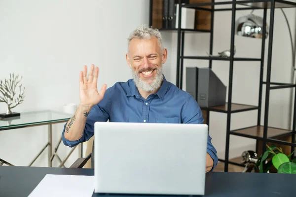 Allegro contemporaneo anziano uomo d'affari capelli grigi si siede di fronte al computer portatile e agitando ciao — Foto Stock