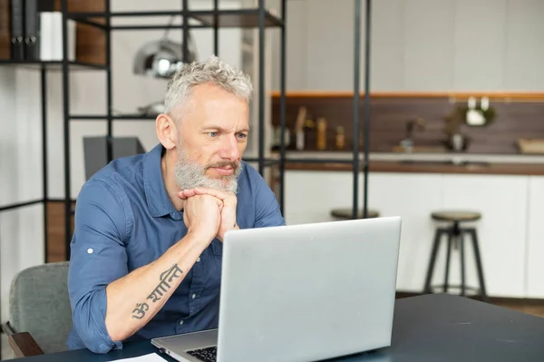 Seriöser Mann mittleren Alters in schickem lässigem Hemd sitzt gedankenverloren mit Laptop am Schreibtisch — Stockfoto
