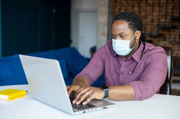 Concentrated African-American male employee wearing medical mask sitting at the desk