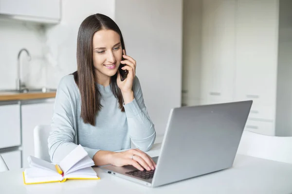 Улыбающаяся и позитивная молодая женщина говорит на смартфоне, работающем с ноутбуком — стоковое фото