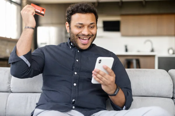 Homem índio sorridente detém cartão bancário e smartphone — Fotografia de Stock