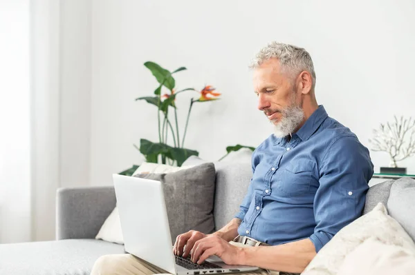 Moderno uomo barbuto di mezza età che utilizza il computer portatile seduto sul divano a casa — Foto Stock