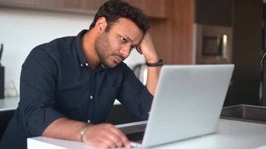 Çok ırklı bir adam evde tek başına otururken dizüstü bilgisayar kullanıyor.