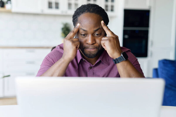 Concentrated African male freelancer using laptop for remote work at home