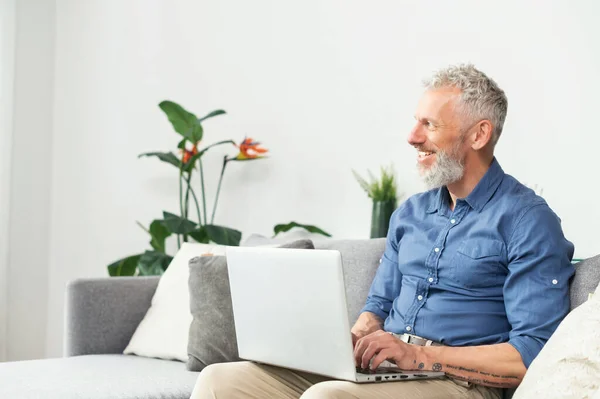 Nowoczesny brodaty mężczyzna w średnim wieku korzystający z laptopa siedzącego na kanapie w domu — Zdjęcie stockowe