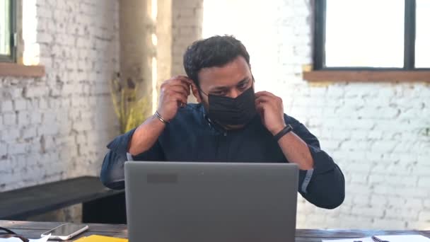 Indian man puts on protective facial mask sitting on the workplace in the office — Stock Video