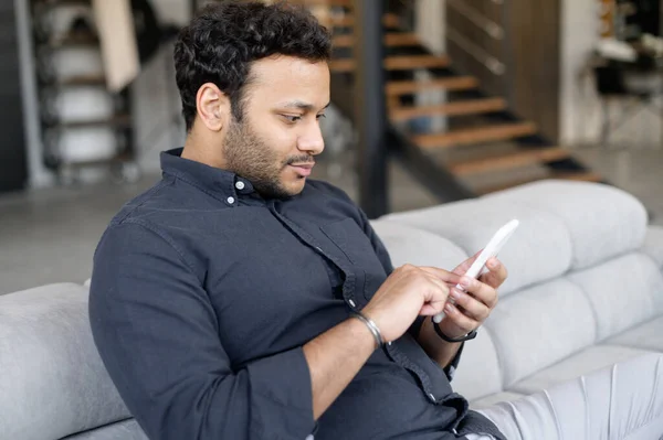 Vista lateral indio joven hombre de negocios usando teléfono inteligente sentado en el sofá en casa —  Fotos de Stock