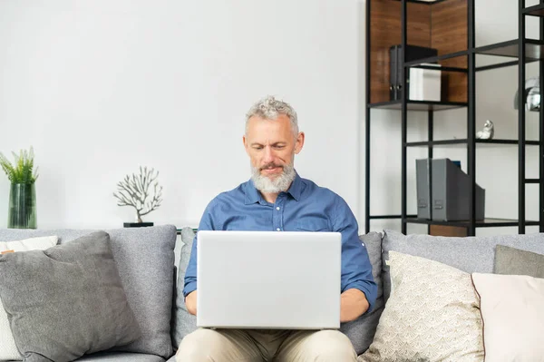 Σύγχρονη μεσήλικας γενειοφόρος άντρας που χρησιμοποιεί φορητό υπολογιστή κάθεται στον καναπέ στο σπίτι — Φωτογραφία Αρχείου