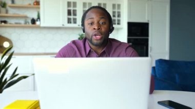 Ciddi bir Afro-Amerikalı erkek. Kulaklık takıyor. Video araması için laptop kullanıyor.