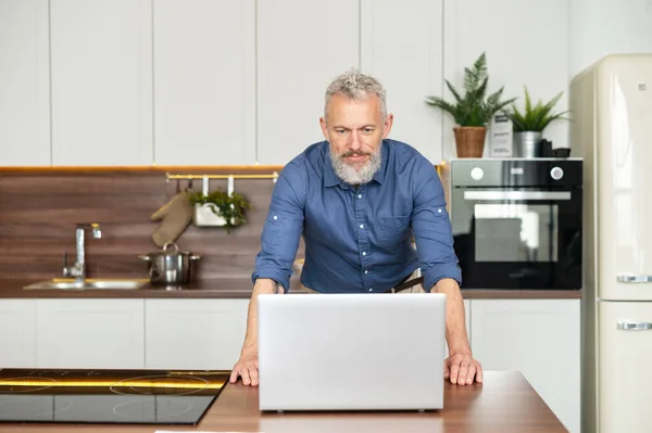 Senior uomo freelance si trova vicino controsoffitto in cucina e fissando il computer portatile — Foto Stock