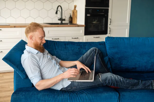 Knappe roodharige man met baard liggend op de bank en met behulp van laptop — Stockfoto