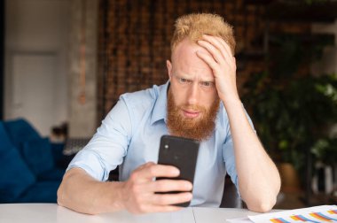 Şaşkın kızıl saçlı iş adamı, şık, rahat gömlekli, akıllı telefon ekranına bakıyor.