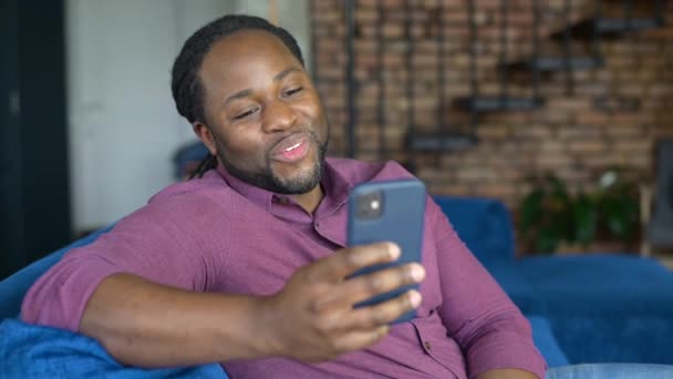 アフリカ系アメリカ人の男を笑顔自宅でソファに座ってスマートフォンを使用して — ストック動画