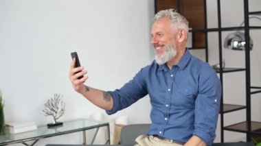 Sıradan giyinen mutlu kıdemli işadamı akıllı telefondan video görüşmesi yapıyor.