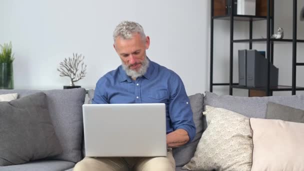 Homem de meia-idade em camisa casual inteligente usando laptop sentado no sofá em casa — Vídeo de Stock