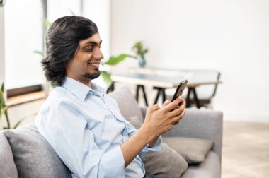 Serene genç Hintli adam akıllı telefonu evdeki kanepede otururken tutuyor.