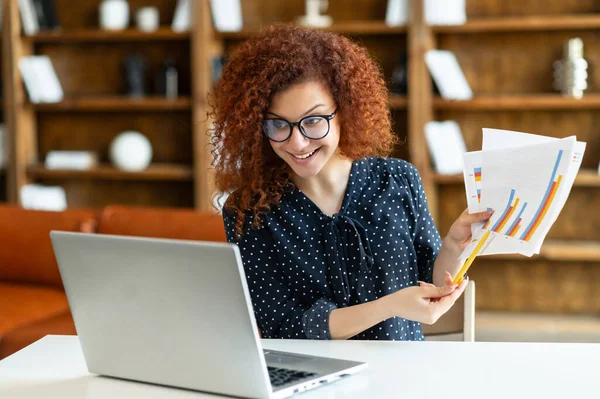 Empleada de oficina sentada en el escritorio de la oficina delante de la computadora portátil y mostrando la hoja con el gráfico —  Fotos de Stock