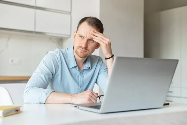 Worried young man using laptop computer for looking for the job, unemployed guy sits in the kitchen at home pondered about future, pensive and upset male job seeker — Stock Photo, Image