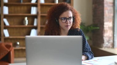 Kablosuz kulaklık takan genç kıvırcık kadın iletişim için dizüstü bilgisayar kullanıyor.