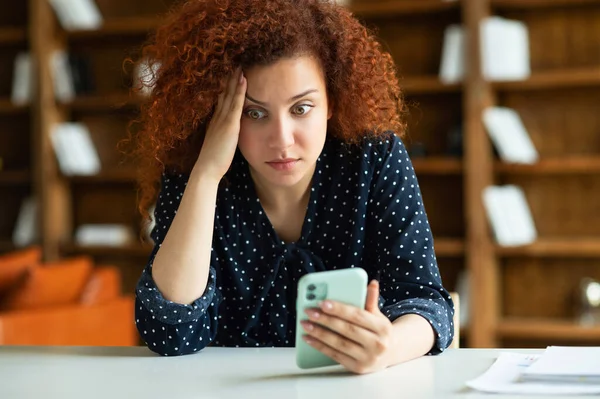 Choqué jeune femme rousse assise au bureau, tenant smartphone — Photo