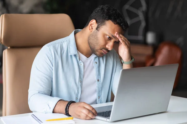 Frustrated sad indian guy is watching at laptop screen and holding head with hand — Stock Photo, Image